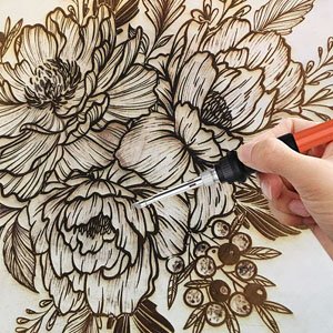 Hand using a wood burning tool to create floral designs on unfinished wood circles