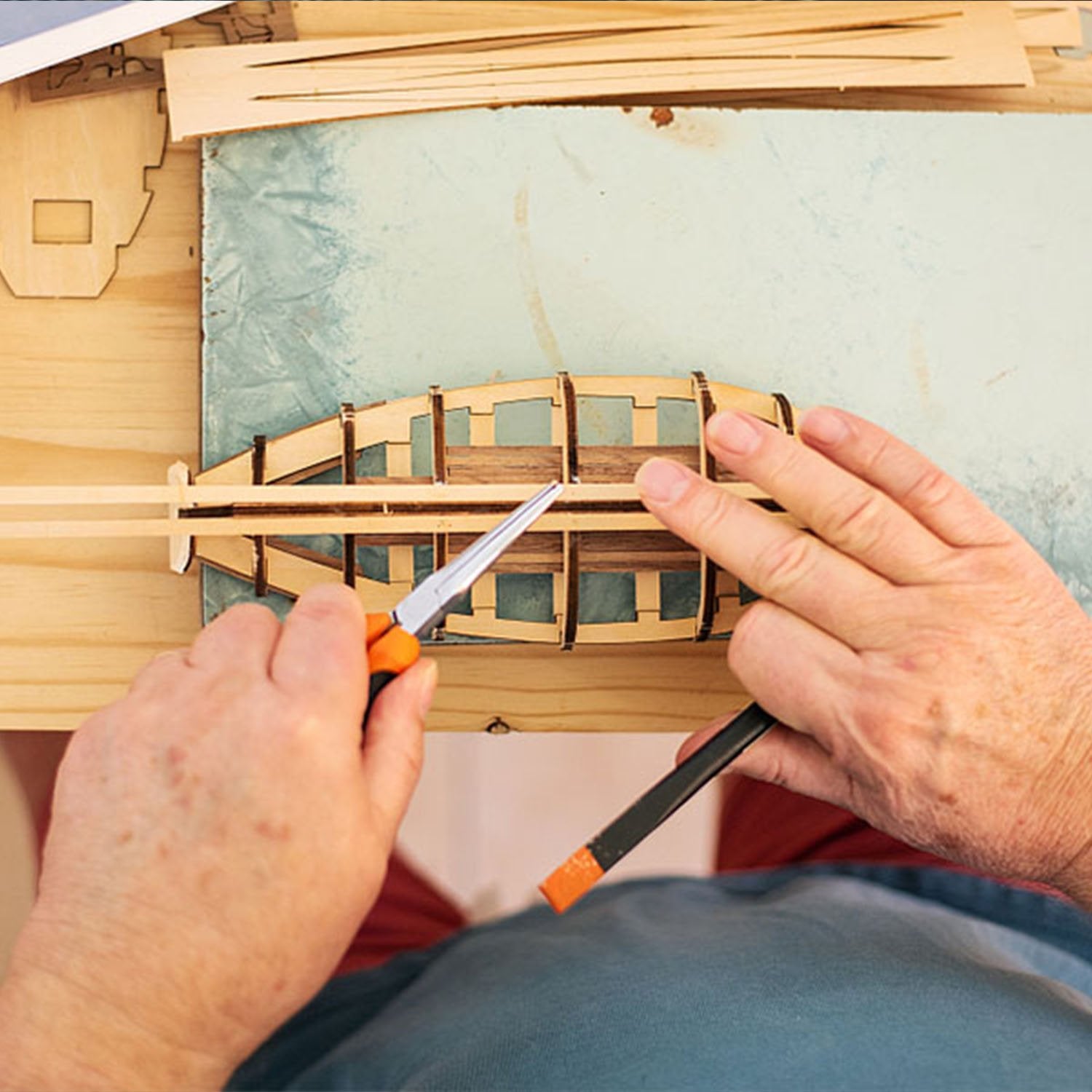 Hands assembling a wooden model kit with tools, close-up overhead view.