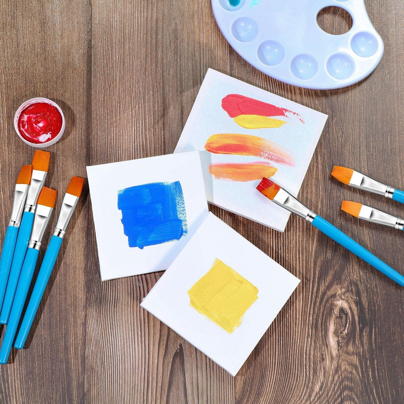 Flat paint brushes with blue handles and orange bristles arranged with small canvases and a paint palette on a wooden surface.