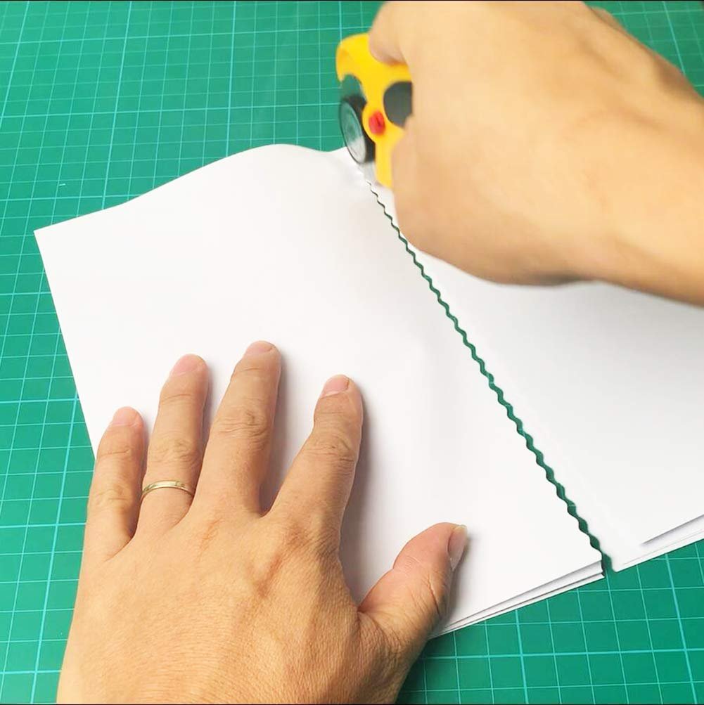 Hands using a yellow pinking rotary cutter to cut a zig-zag edge on white paper on a green cutting mat.