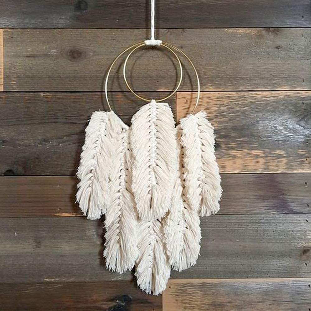 Macrame wall hanging with two gold metal rings and cream-colored feather-like tassels, suspended against a wooden plank background.