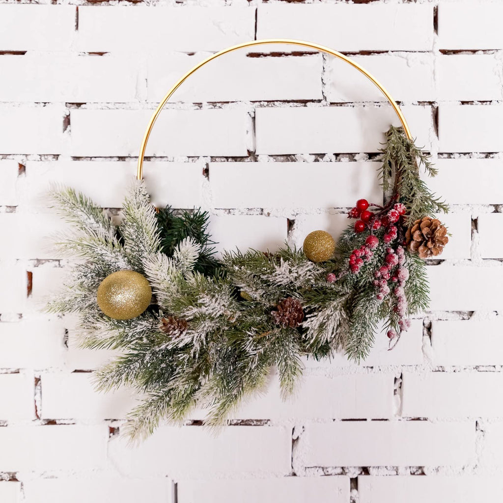 Gold metal ring wreath decorated with frosted pine branches, red berries, pinecones, and gold ornaments against a white brick wall.