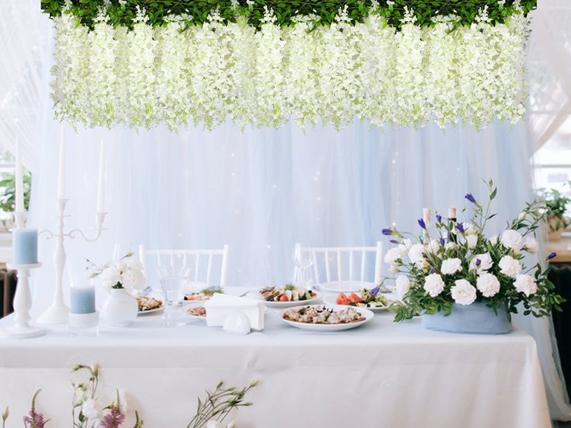 White artificial wisteria garlands draped above a decorated table setting with food and flowers.