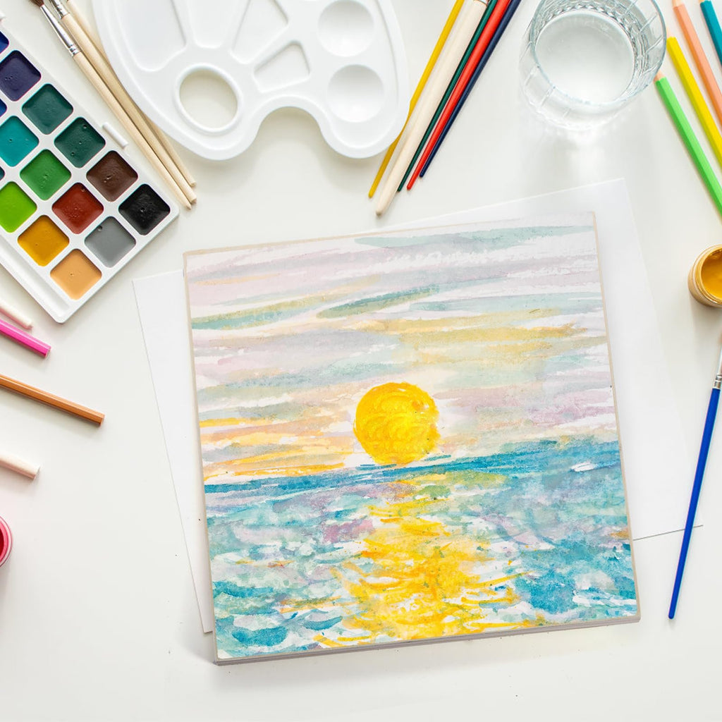 Watercolor painting of a sunset over the ocean, with art supplies including a palette, brushes, and colored pencils arranged around it.