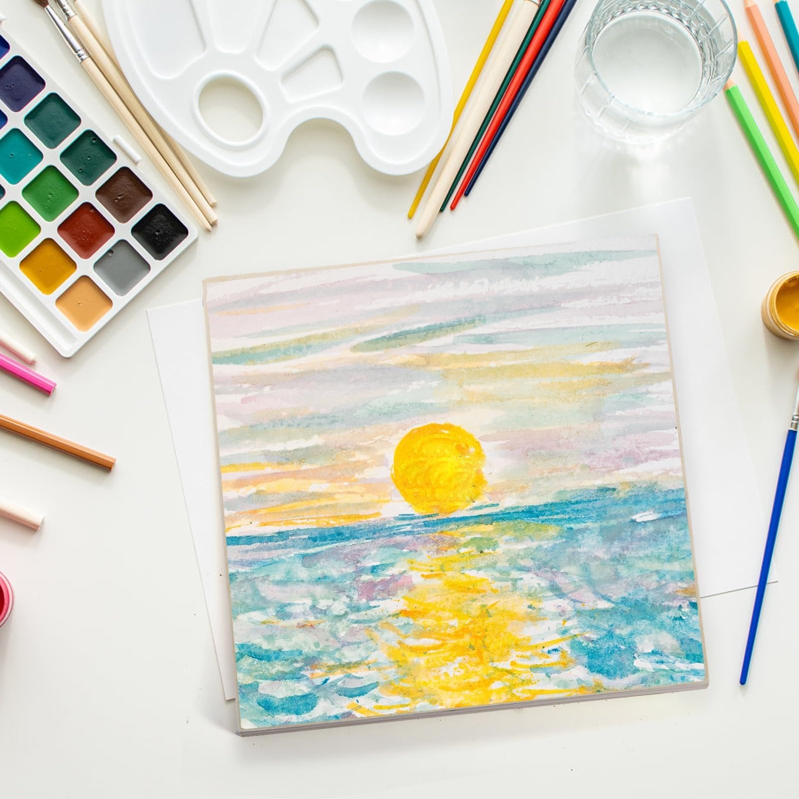 Watercolor painting of a sunset over the ocean, with art supplies including a palette, brushes, and colored pencils arranged around it.