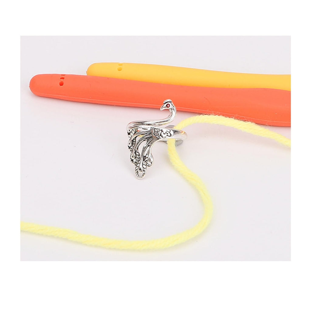 Silver peacock-shaped crochet loop ring with yellow yarn threaded through it, next to orange and yellow crochet hooks.