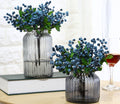 Two vases filled with artificial blueberry stems and green leaves, one vase on a book, a wine glass to the right.