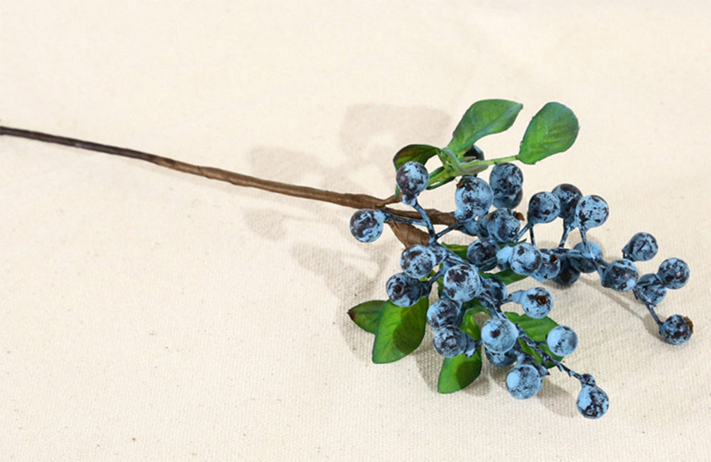 Artificial blueberry stem with blue berries and green leaves on a textured off-white background.