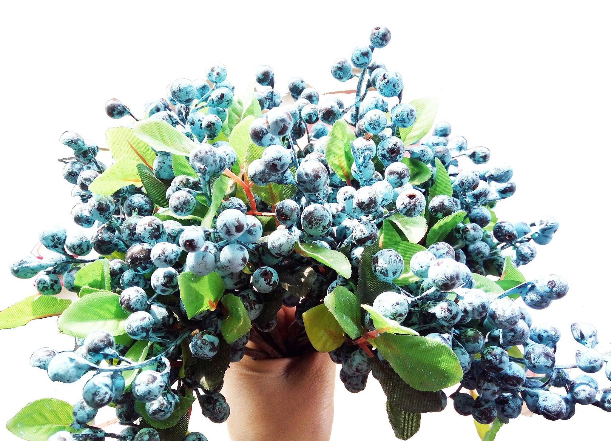 Close-up of artificial blueberry stems with green leaves, held by a hand against a white background.