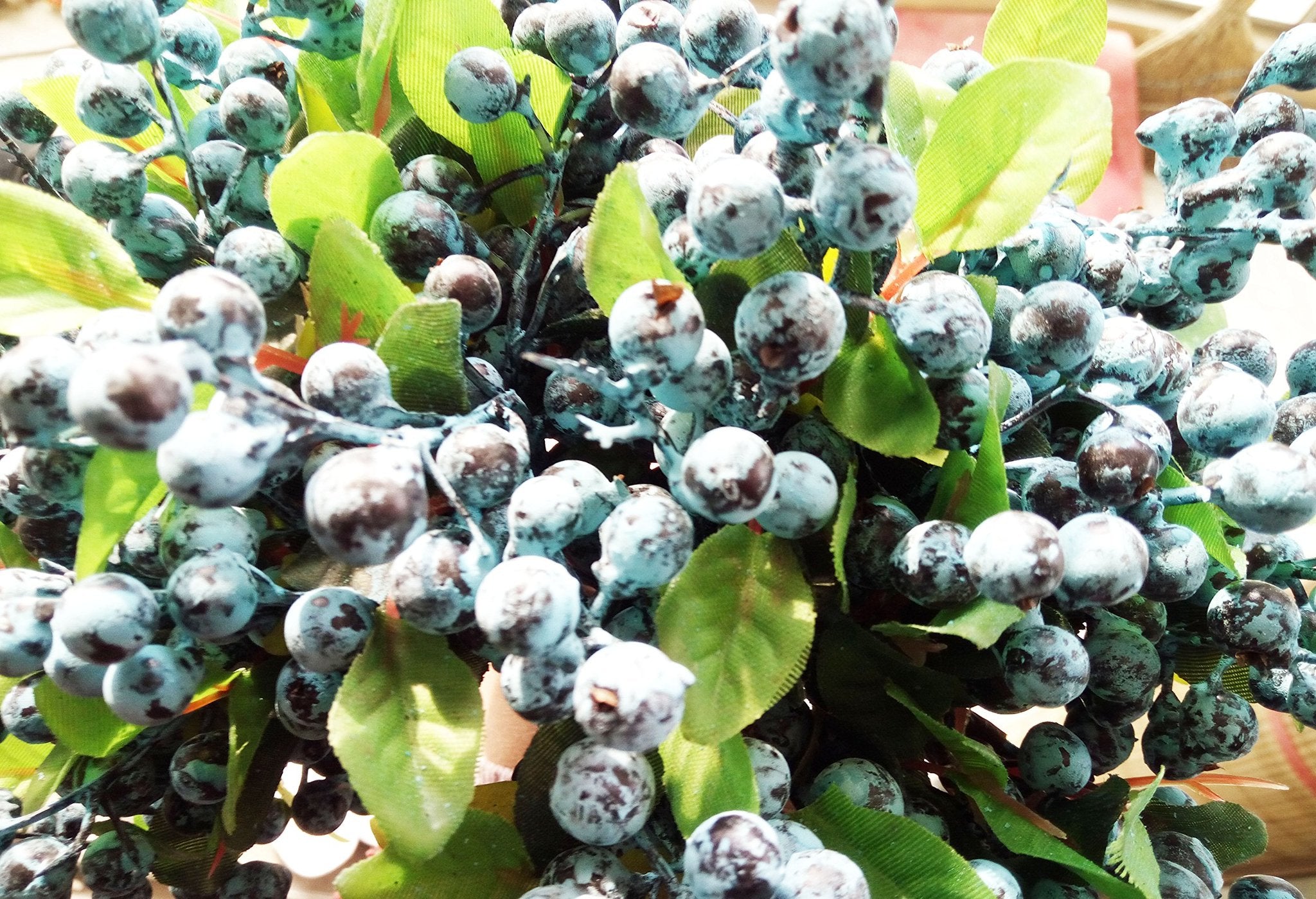 Close-up of artificial blueberry stems with green leaves, showing clusters of blue berries with dark speckles.