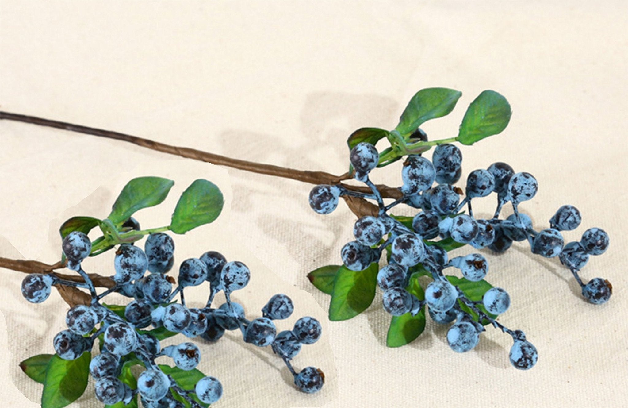 Two artificial blueberry stems with green leaves and blue berries on a textured off-white background.