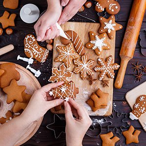 Hands decorating gingerbread cookies with white icing on a wooden cutting board, surrounded by cookie cutters and ingredients.