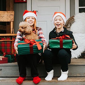 Two children wearing Santa hats and holding wrapped gifts and a dog on a porch during Christmas.