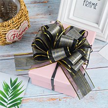 Pink gift box with a large black and gold pull bow on top, next to a basket and a white frame.