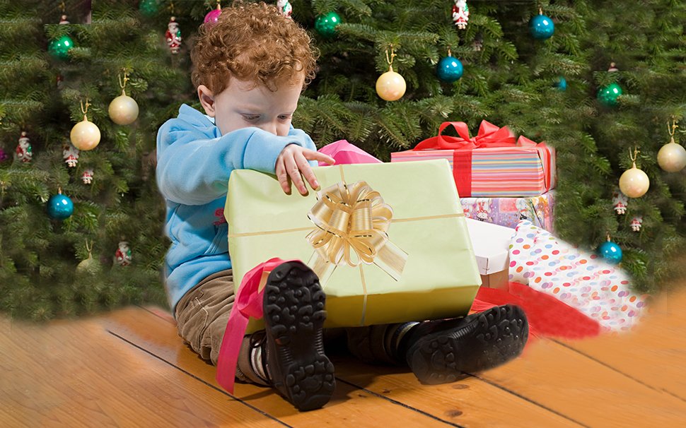 Young child sitting on wooden floor opening a large gift box with a gold bow, near a decorated Christmas tree.