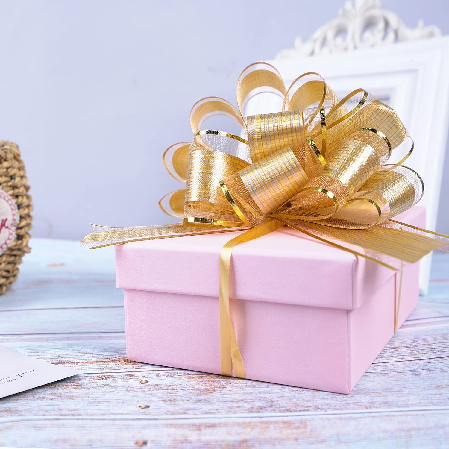 Pink gift box with a large gold pull bow on top, set on a wooden surface with a decorative frame in the background.