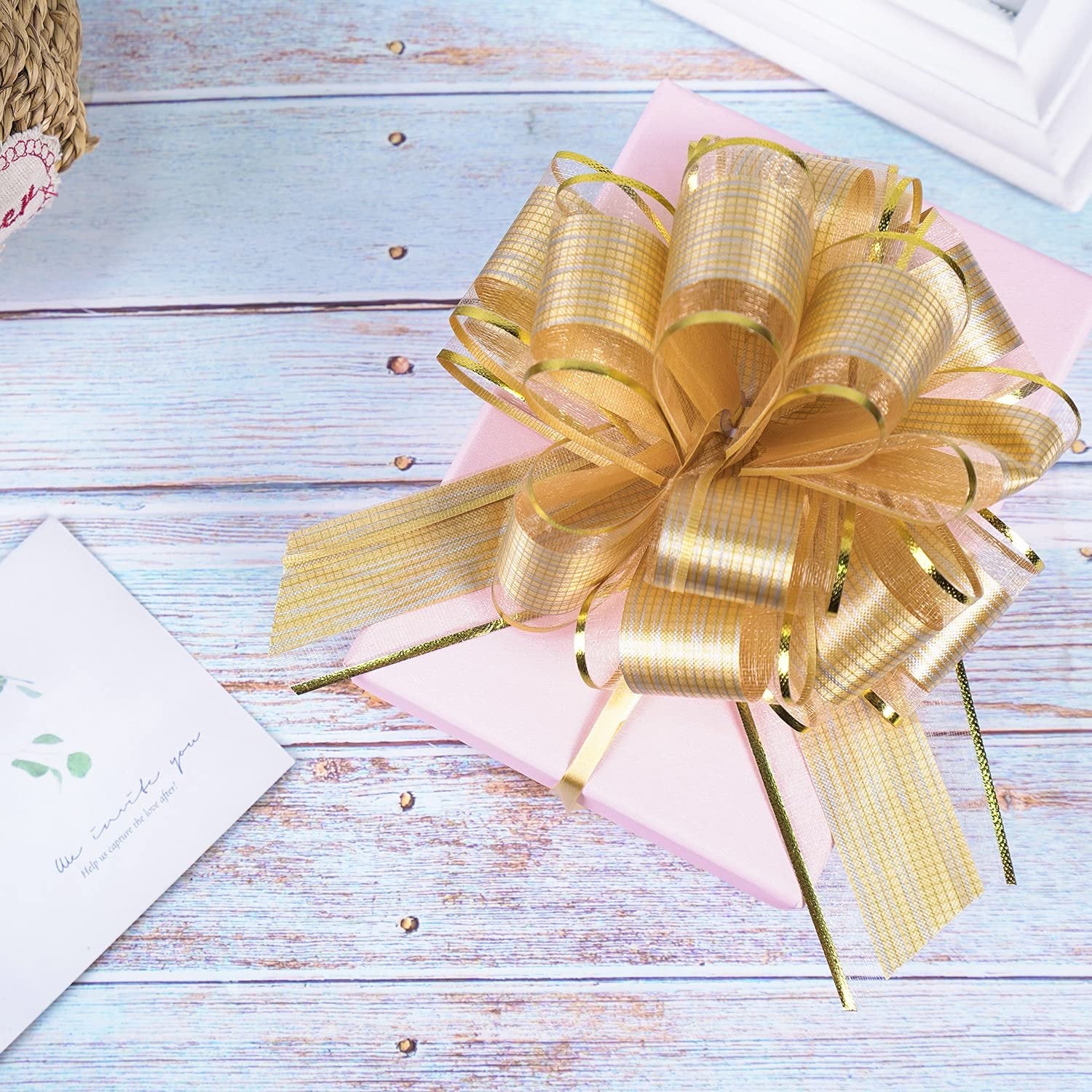 Pink gift box with a large gold pull bow on a light blue wooden surface, next to a card.