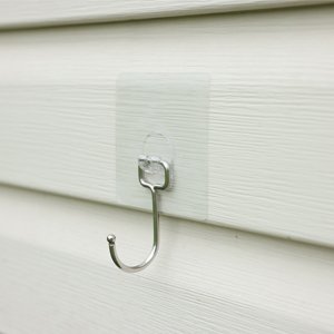 Large transparent adhesive wall hook attached to white siding, showing metal hook detail.