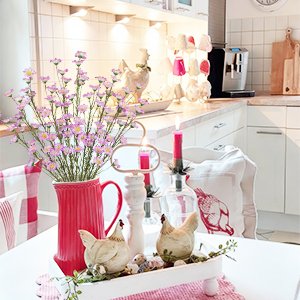 Pink forsythia branches in a red pitcher on a table with decorative chickens and a candle.