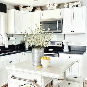 Kitchen island with a vase of artificial yellow and white forsythia branches, lemons in a bowl, and stools in a bright white kitchen.