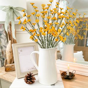 Ten artificial yellow forsythia branches in a white pitcher on a table with a pinecone and candle.
