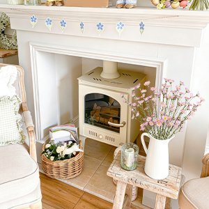 Pink forsythia branches in a white pitcher on a wooden stool next to a fireplace with an electric stove.