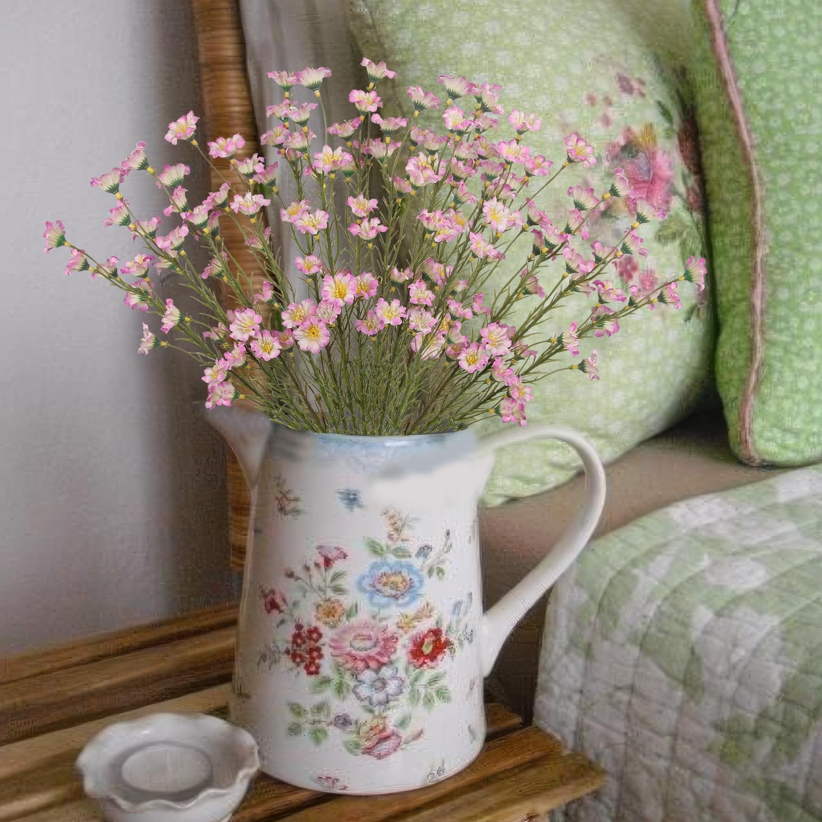 Artificial pink forsythia branches in a floral pitcher next to green pillows and a quilt.