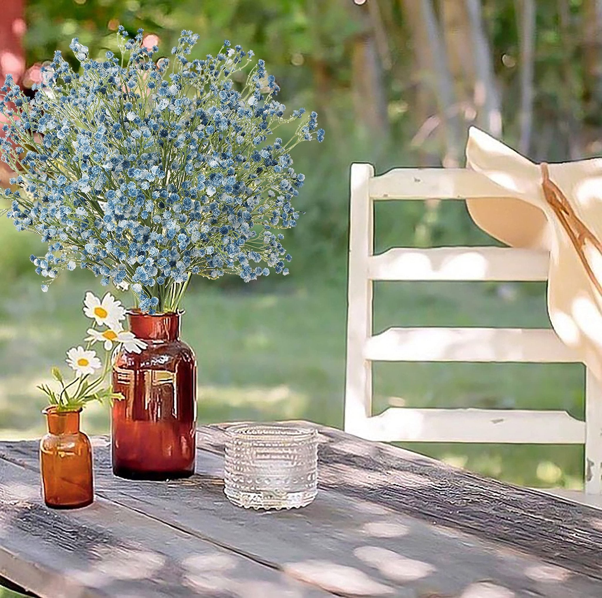 Bouquet of blue artificial baby's breath flowers in amber vases on a wooden table with a chair and hat in the background.