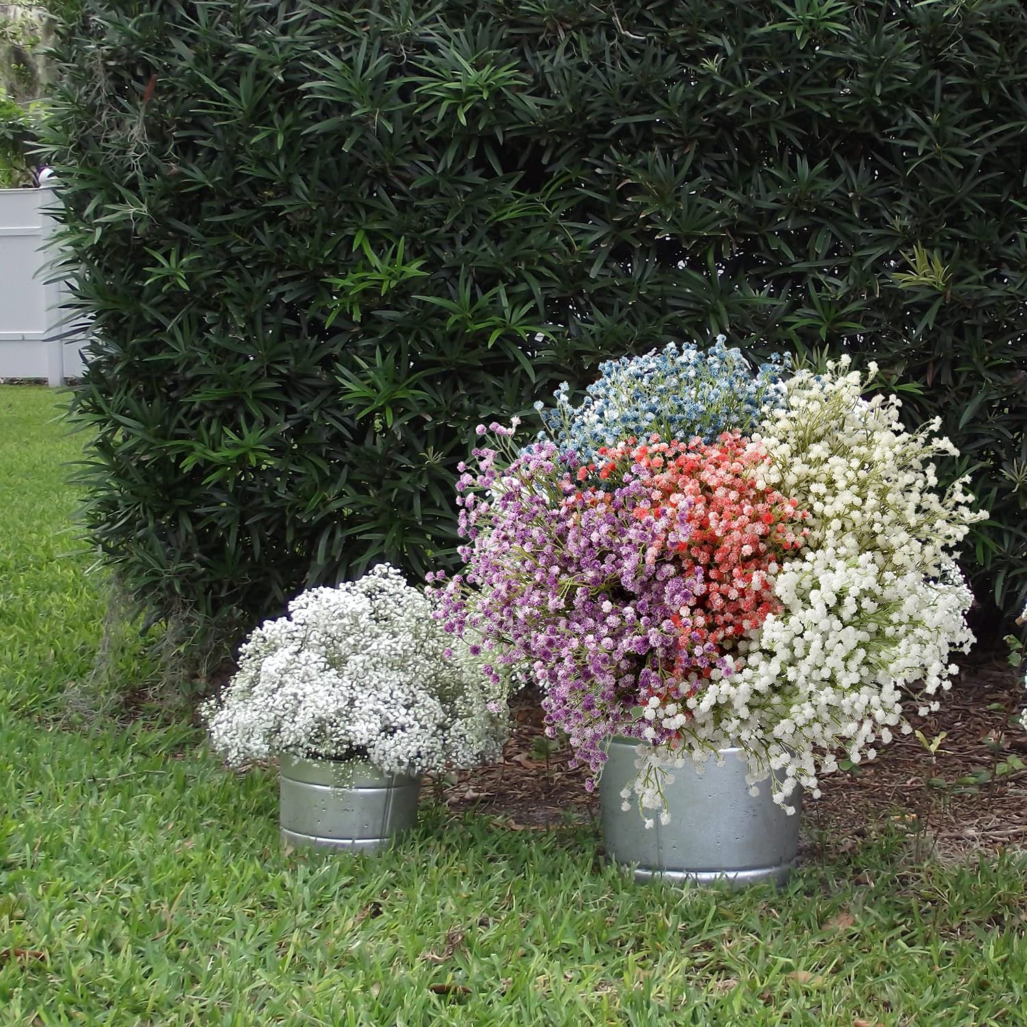 Bunches of artificial baby's breath flowers in various colors displayed in galvanized buckets on grass with a green hedge background.