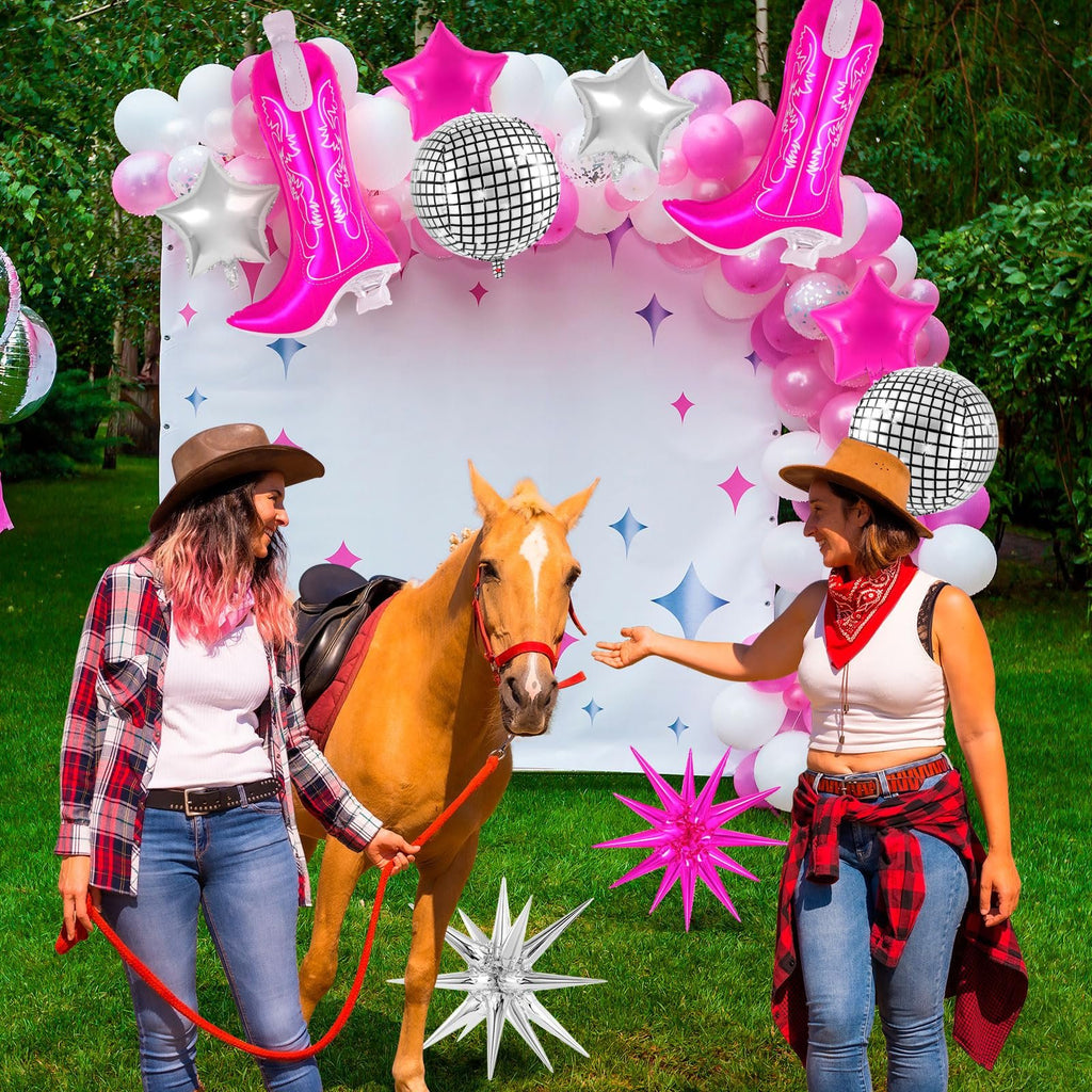 Two women in cowboy hats with a horse in front of a balloon arch featuring pink cowgirl boot balloons and disco balls.