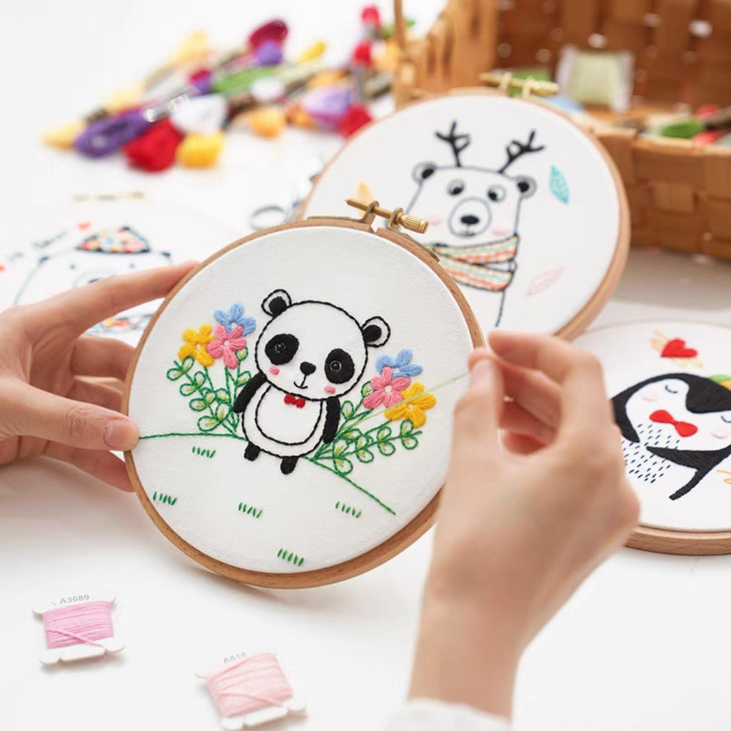Hands embroidering a panda design on white fabric in a wooden hoop, with other embroidery hoops and thread spools visible.
