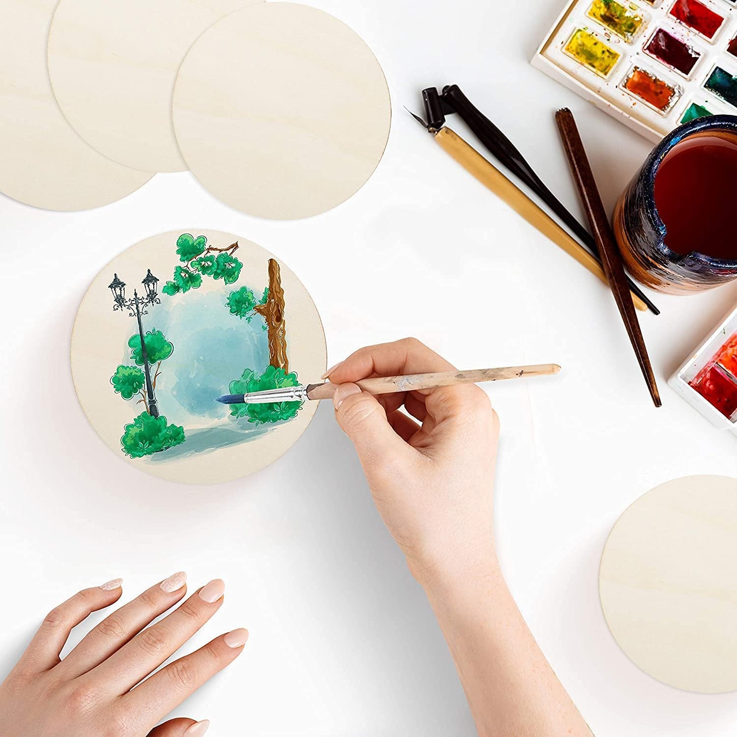 Unfinished wood circles with watercolor painting supplies and a hand painting a park scene on one circle.