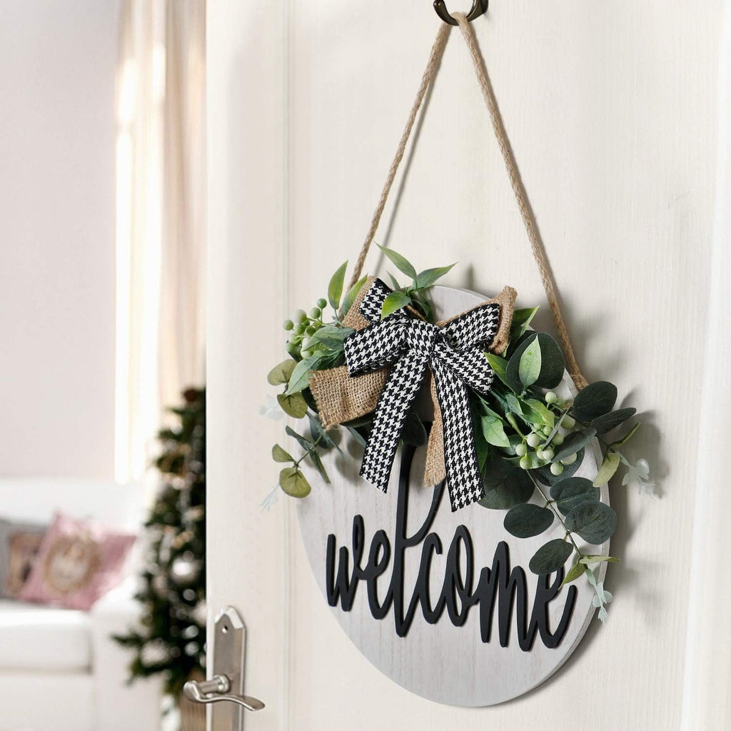 Round wooden welcome sign with faux greenery and a black and white bow hanging on a door.