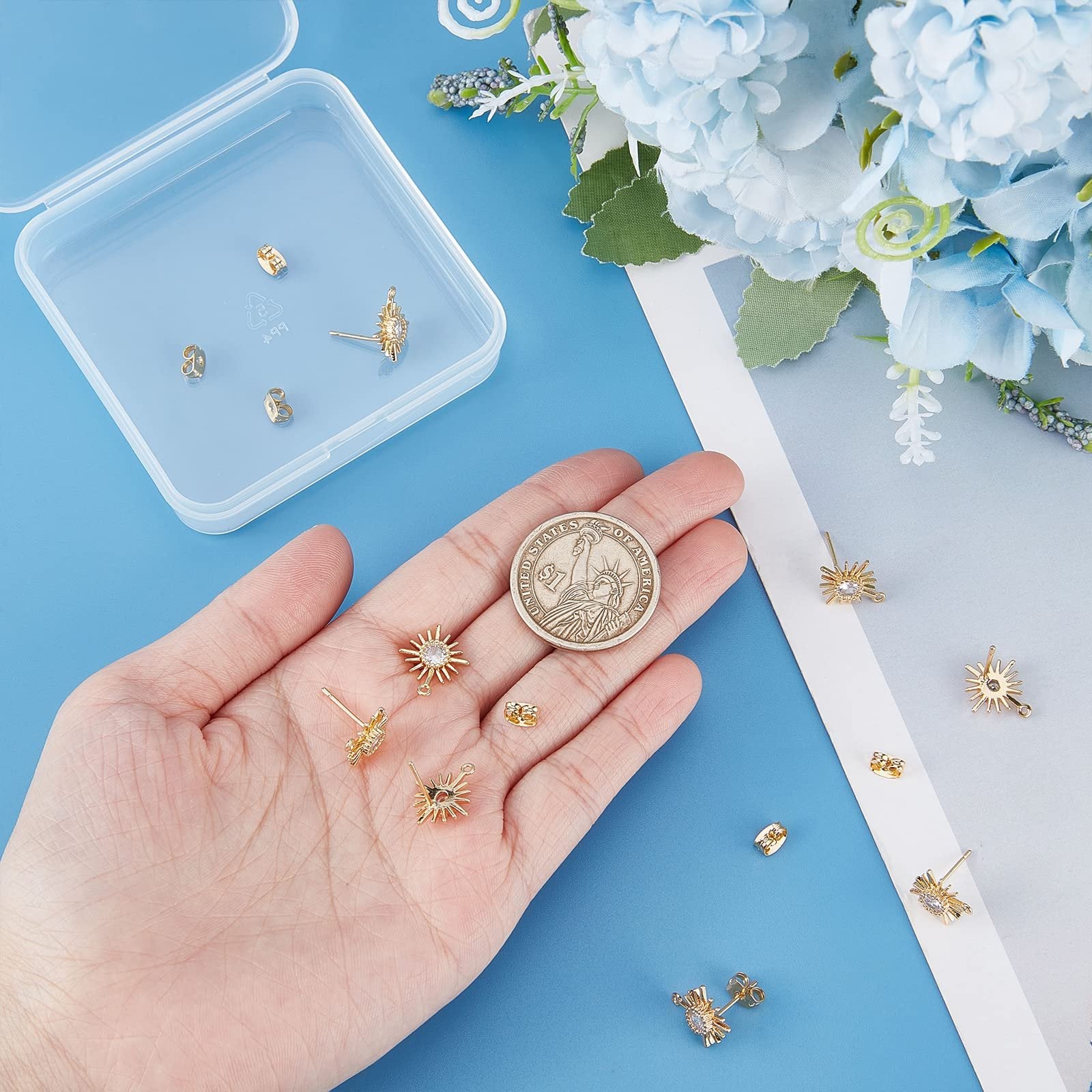 Hand holding gold sun-shaped earring findings with cubic zirconia and a US quarter for scale, with more findings in a clear plastic box and scattered on a blue surface.