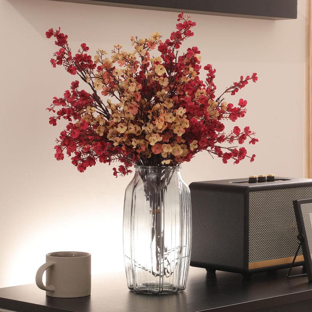 Artificial baby's breath flowers in red and beige hues arranged in a clear glass vase on a dark shelf next to a mug and a speaker.