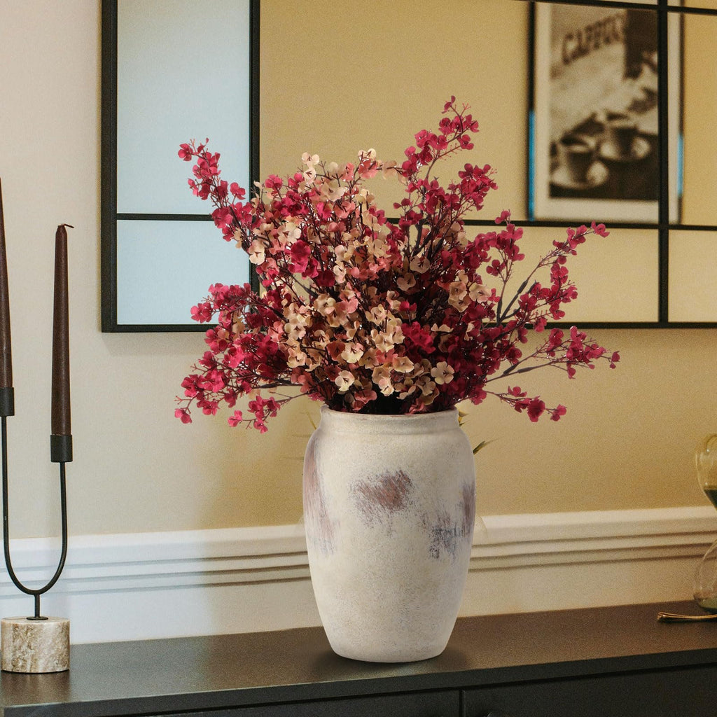 Artificial baby's breath flowers in a distressed vase on a dark surface with candles and a framed picture in the background.