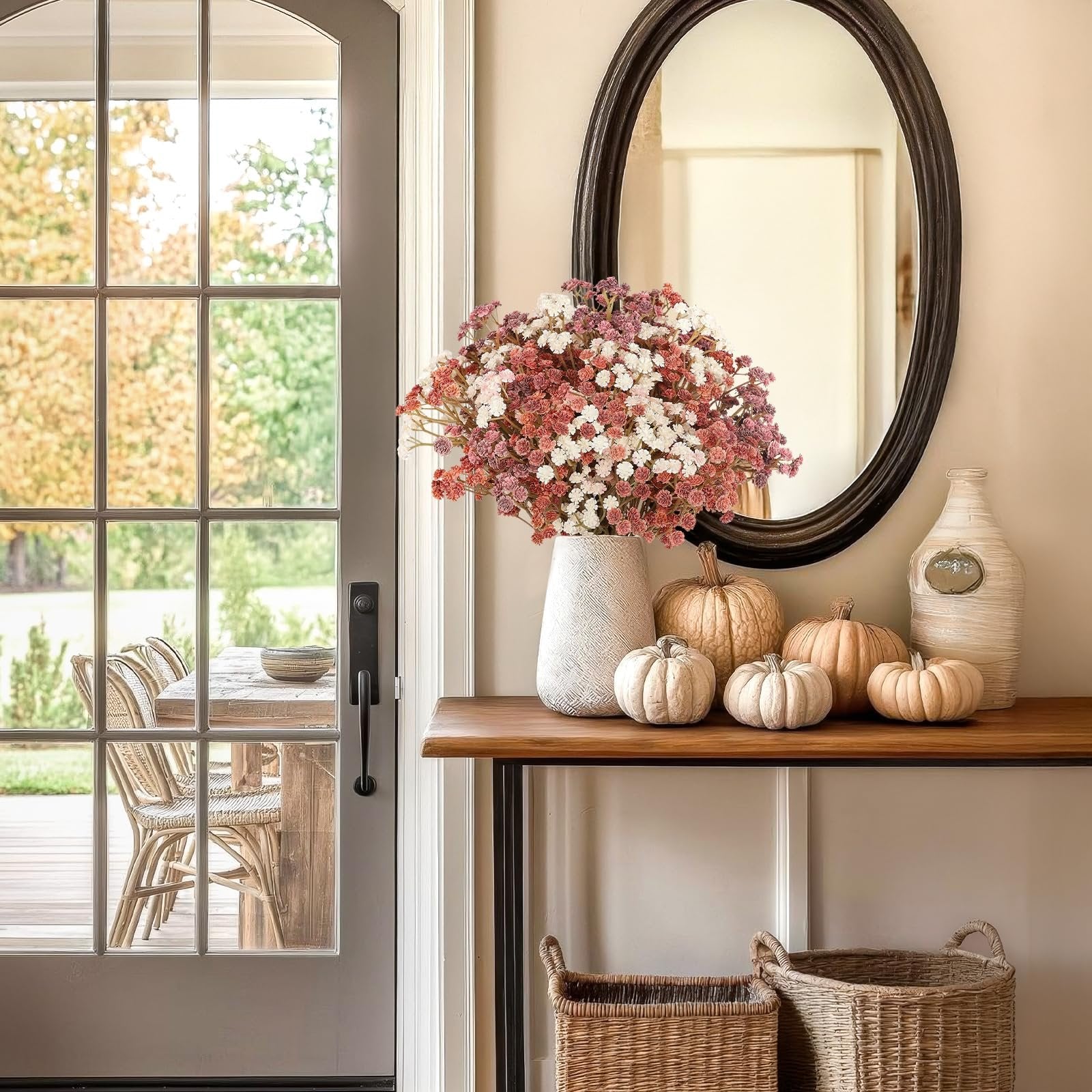 Fall decor arrangement with a vase of pink and white baby's breath flowers, pumpkins, and decorative bottles on a console table.