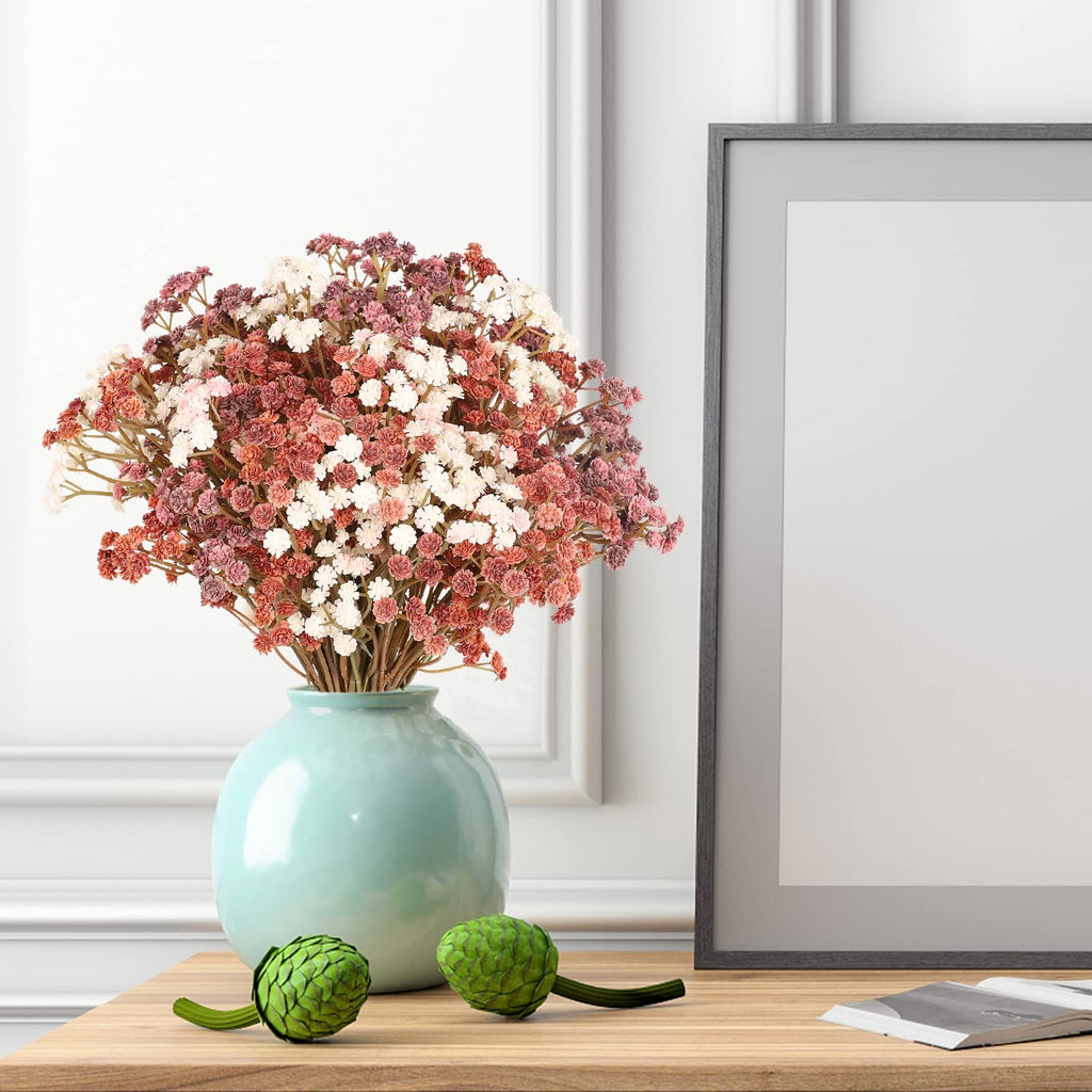 Bouquet of artificial baby's breath flowers in mixed pink, white, and coral hues in a teal vase, next to a picture frame on a wooden surface.
