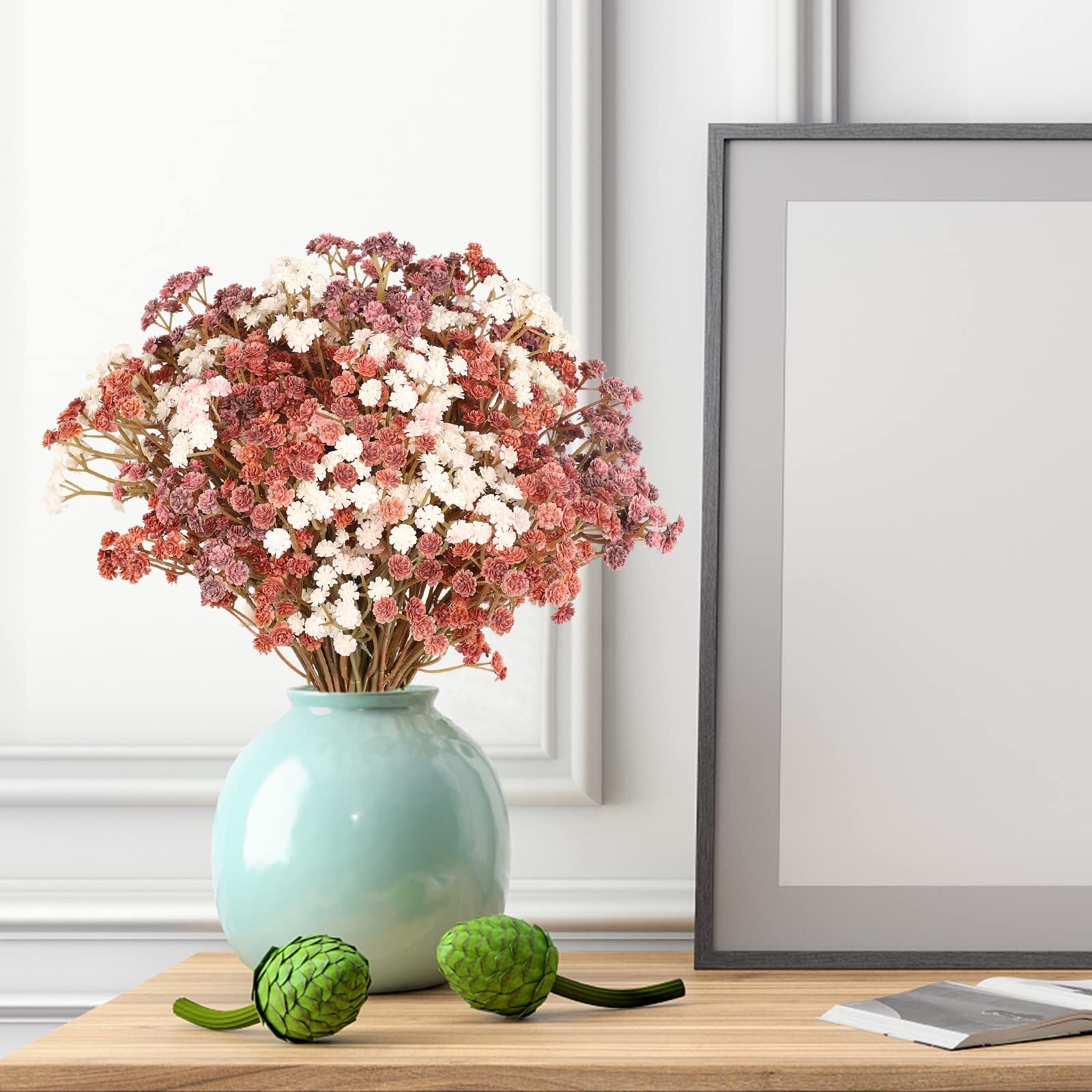 Bouquet of artificial baby's breath flowers in mixed pink, white, and coral hues in a teal vase, next to a picture frame on a wooden surface.