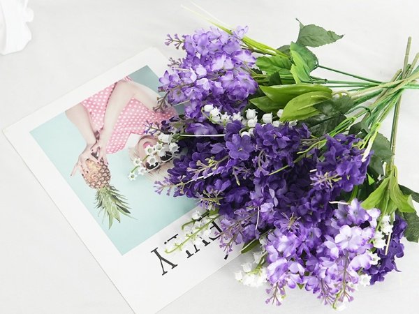 Bouquet of artificial purple lilac and white campanula flowers with green leaves, resting on a magazine with a woman holding a pineapple.