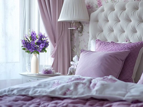 Artificial purple lilac and white campanula flowers in a cream vase on a bedside table.