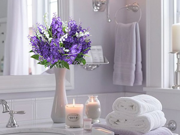 Artificial purple lilac and white campanula flowers in a vase on a bathroom counter with towels and candles