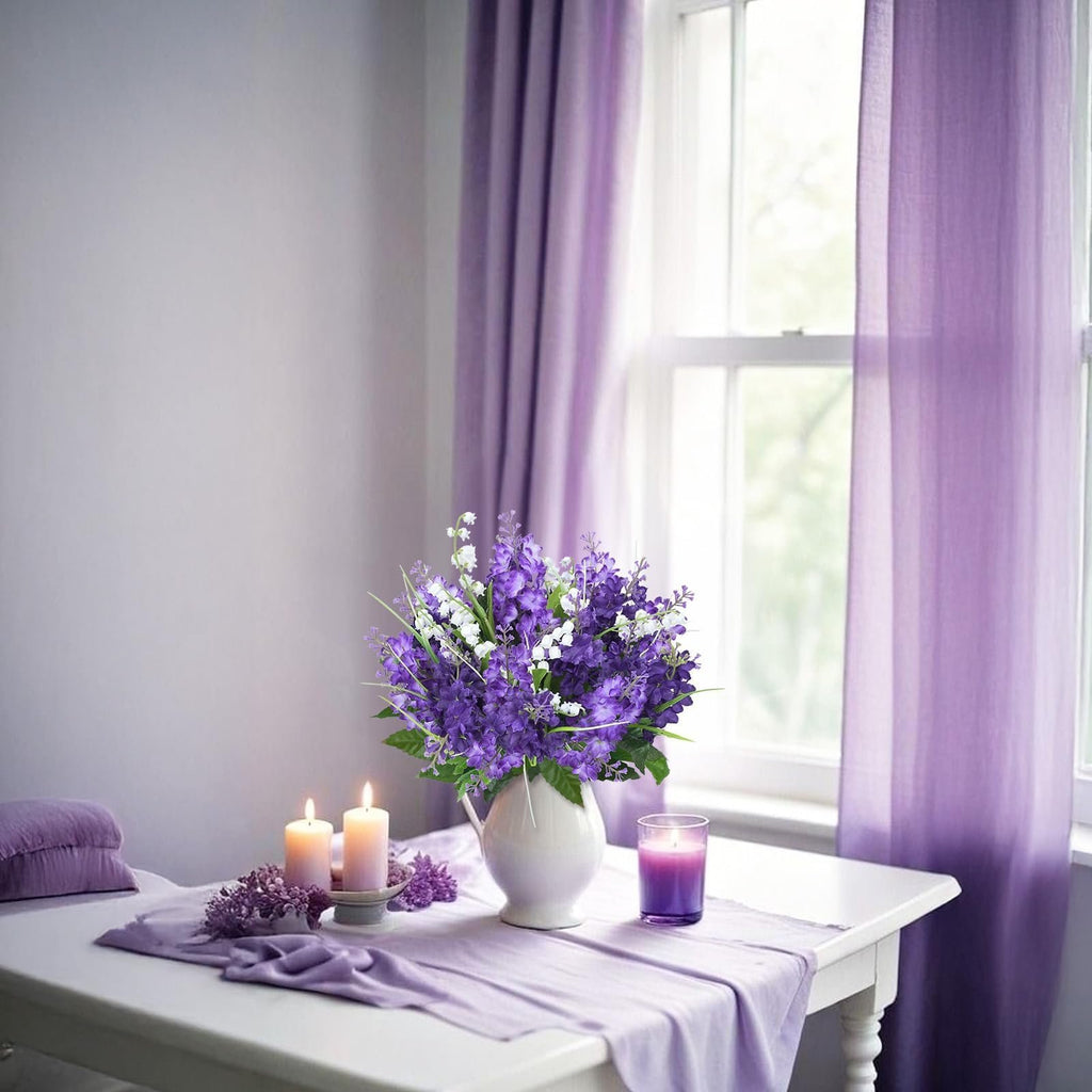 Artificial purple lilac and white campanula flowers in a white vase on a table with lit candles and purple curtains.