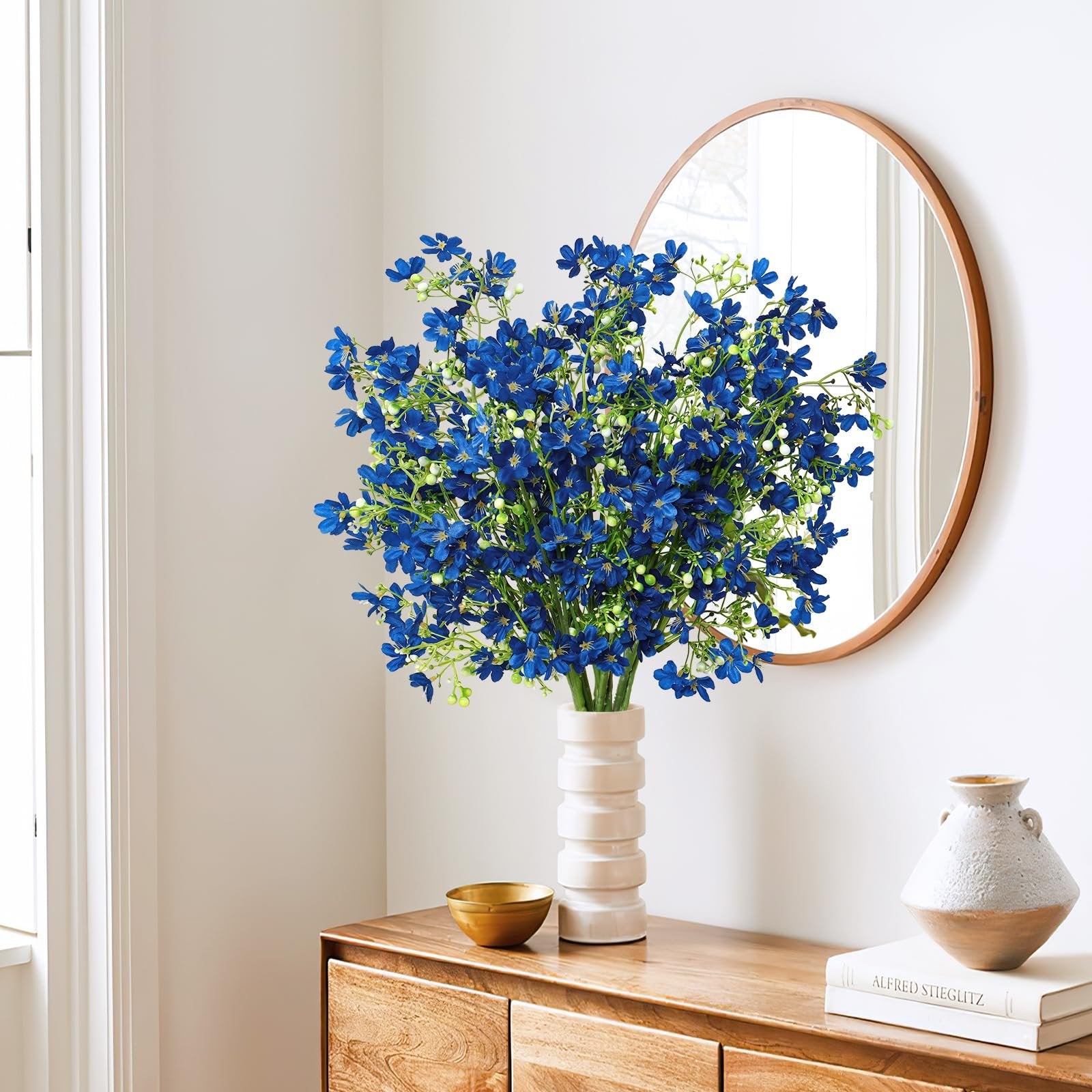 Bouquet of blue silk flowers in a white vase on a wooden dresser, with a round mirror in the background.