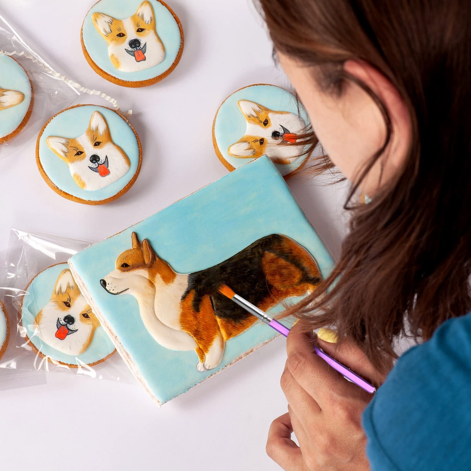 Person decorating a rectangular cake with a corgi design using a purple brush, surrounded by round corgi-themed cookies.