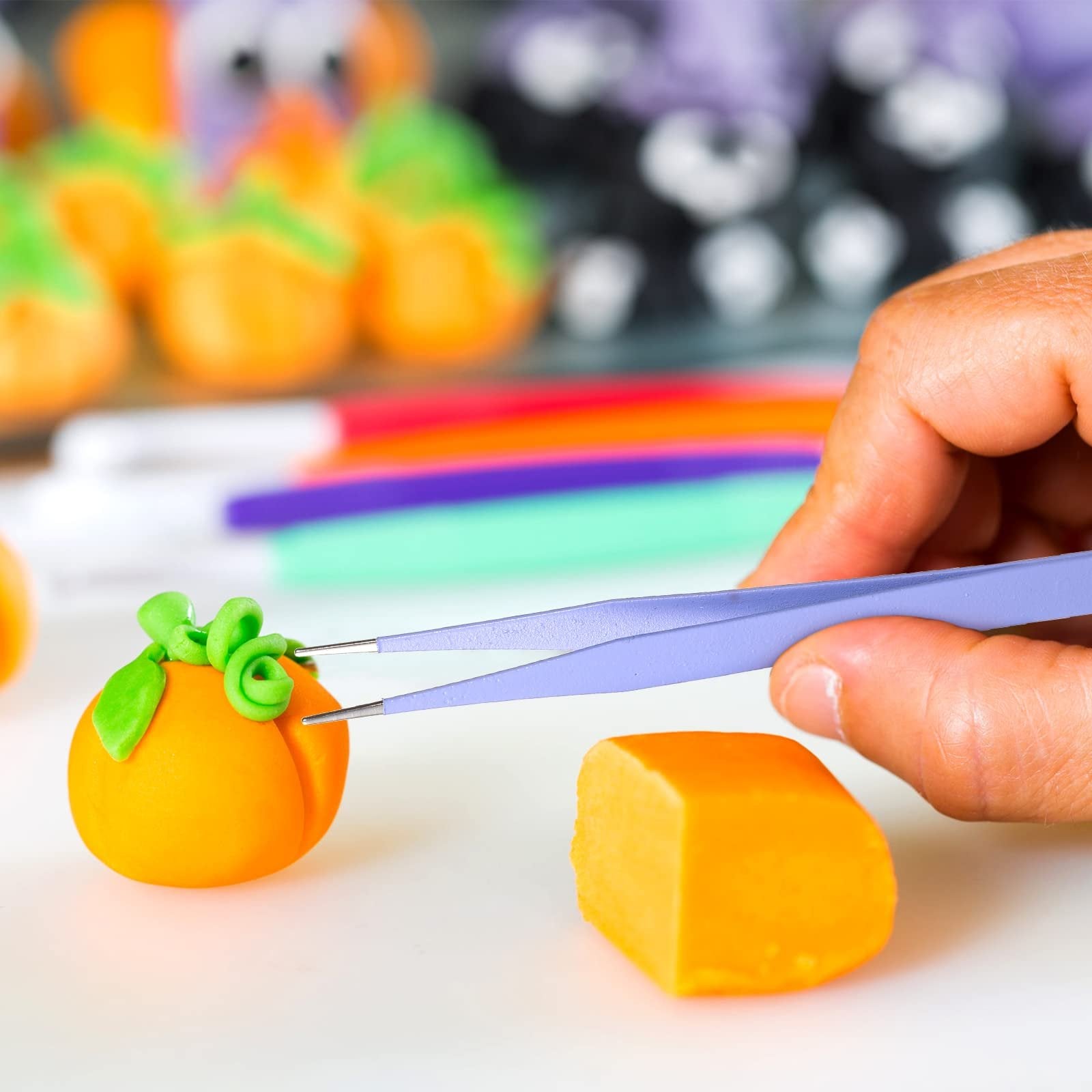 Hand using purple tweezers to decorate an orange fondant pumpkin with green fondant leaves and swirls.