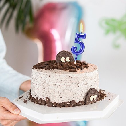 Blue number 5 birthday candle on a cake decorated with cookies and chocolate crumbs, held by a person.