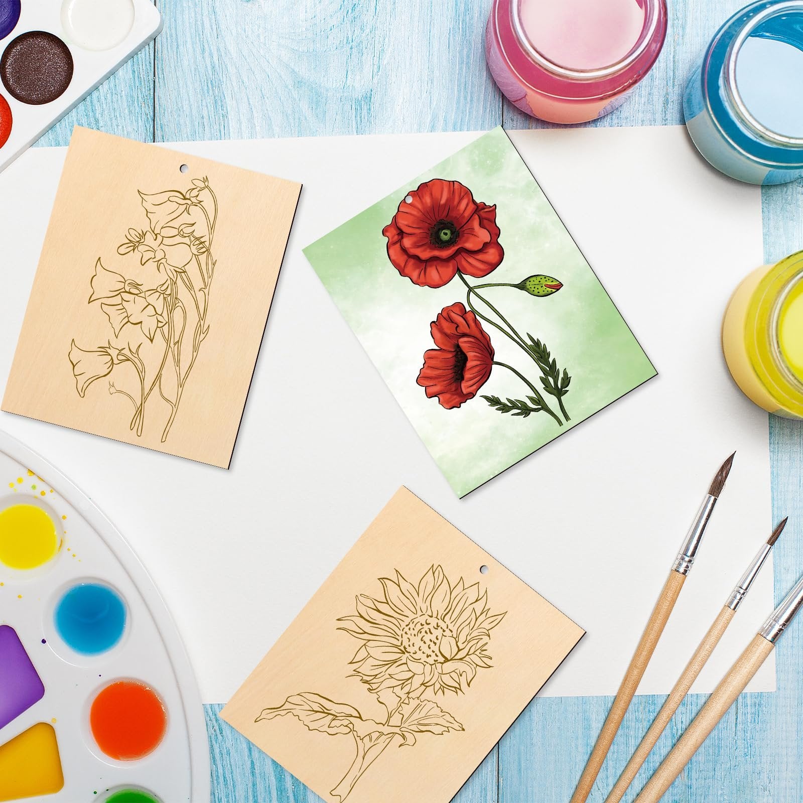 Three unfinished wood ornaments with floral designs, paint palette, brushes, and paint jars on a blue wooden surface.