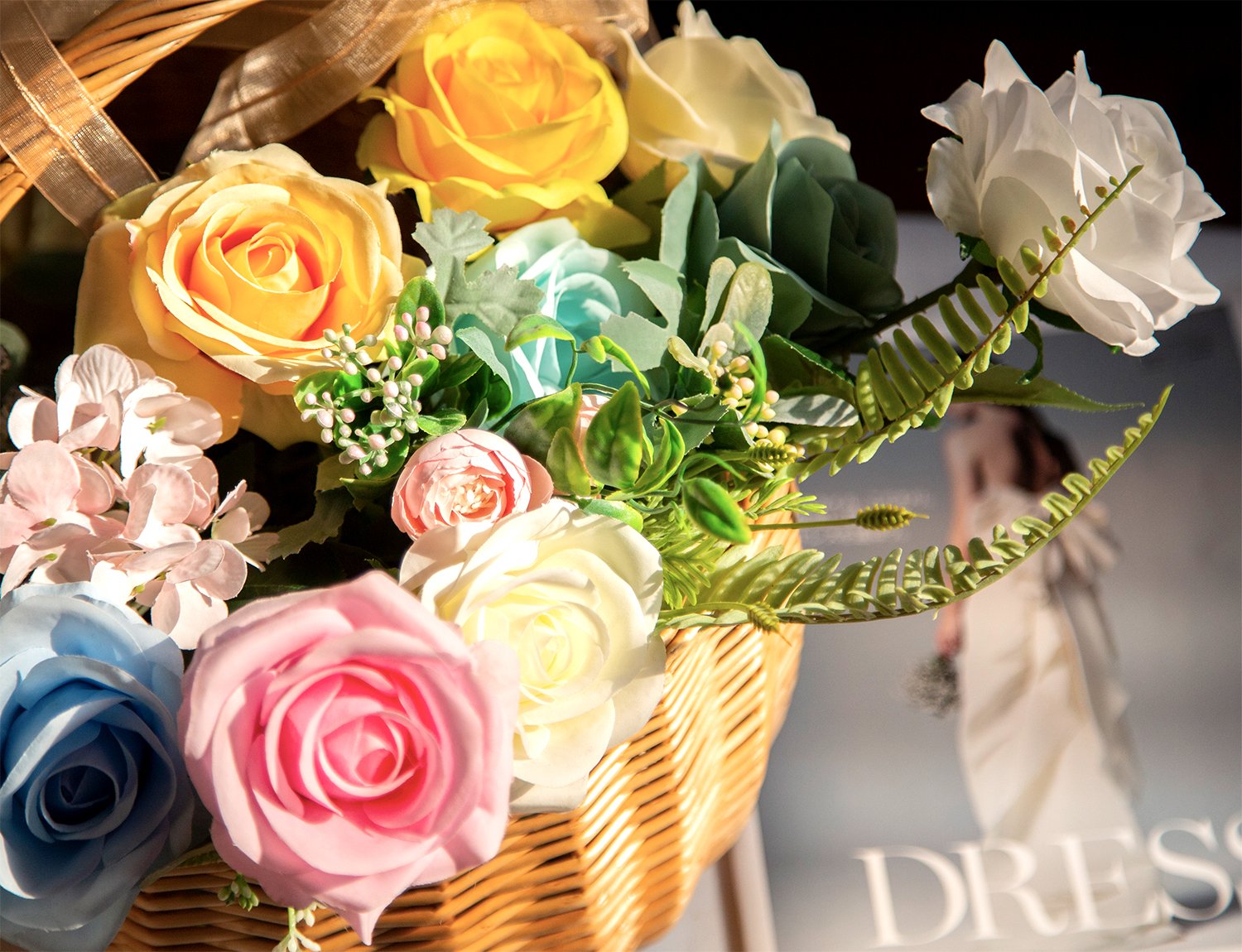 Close-up of a woven basket filled with colorful artificial roses and greenery, with a magazine featuring a wedding dress visible in the background.