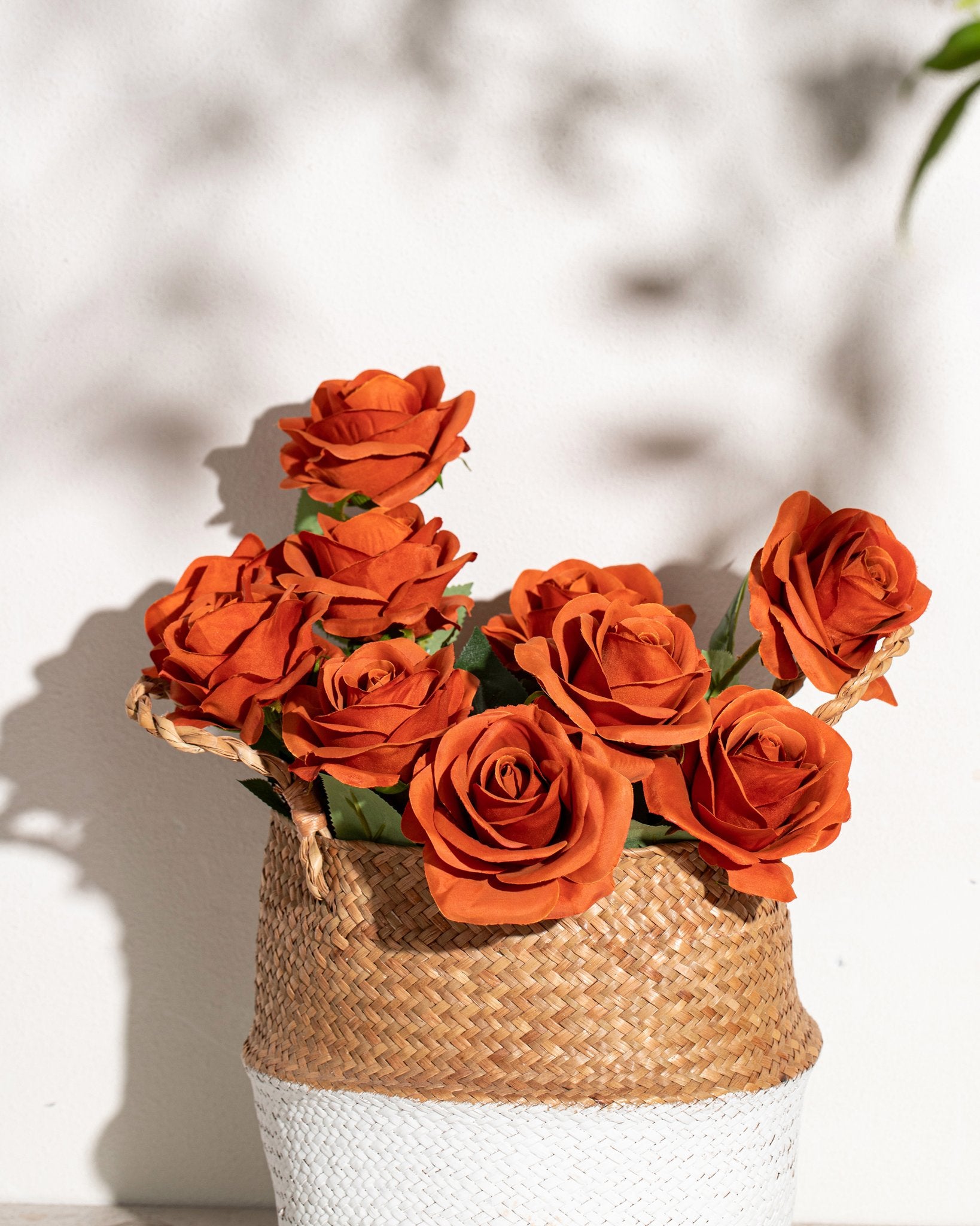 Bouquet of 10 rust-colored artificial roses in a woven basket with white base, against a textured white wall with shadows.